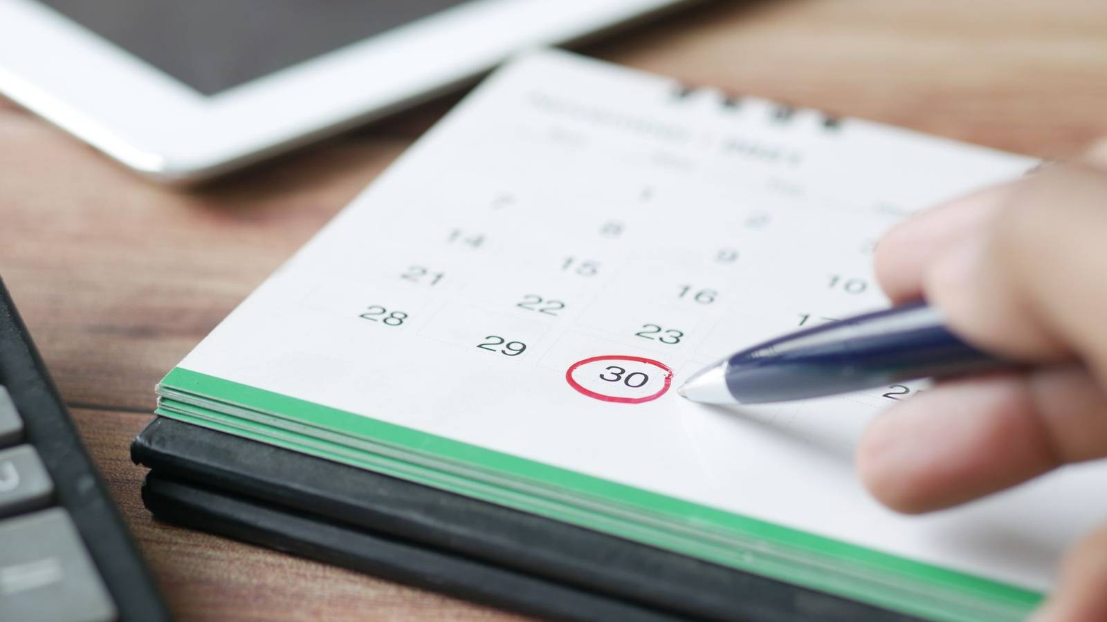 卓上カレンダーの日付に赤い丸が付けられ、ペンを持った手がその横に添えられている様子